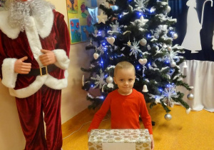 Uśmiechnięty chłopiec trzyma w ręku prezent, stoi obok figury Mikołaja.